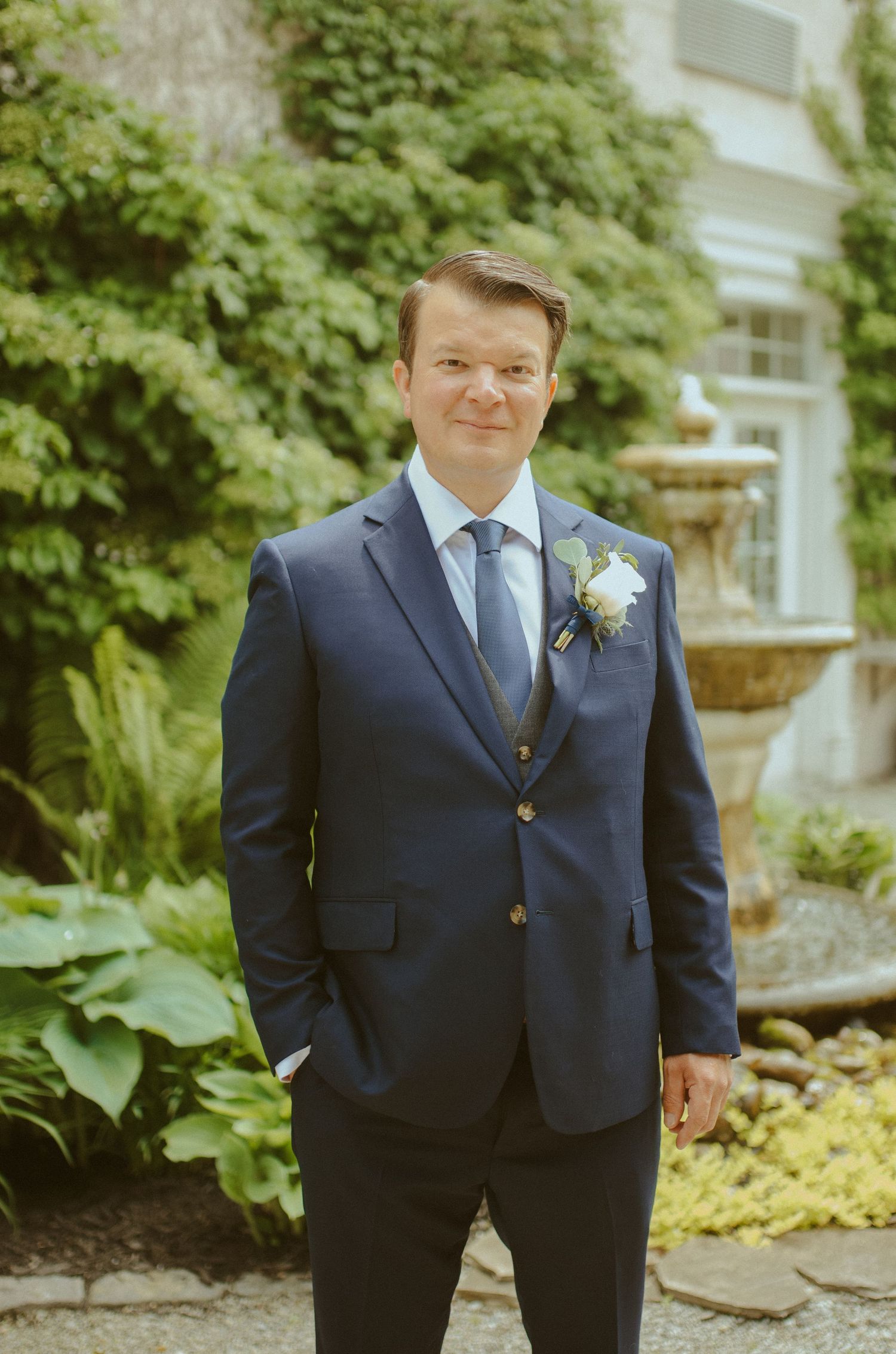 A groom in a classic navy suit poses in a garden setting.