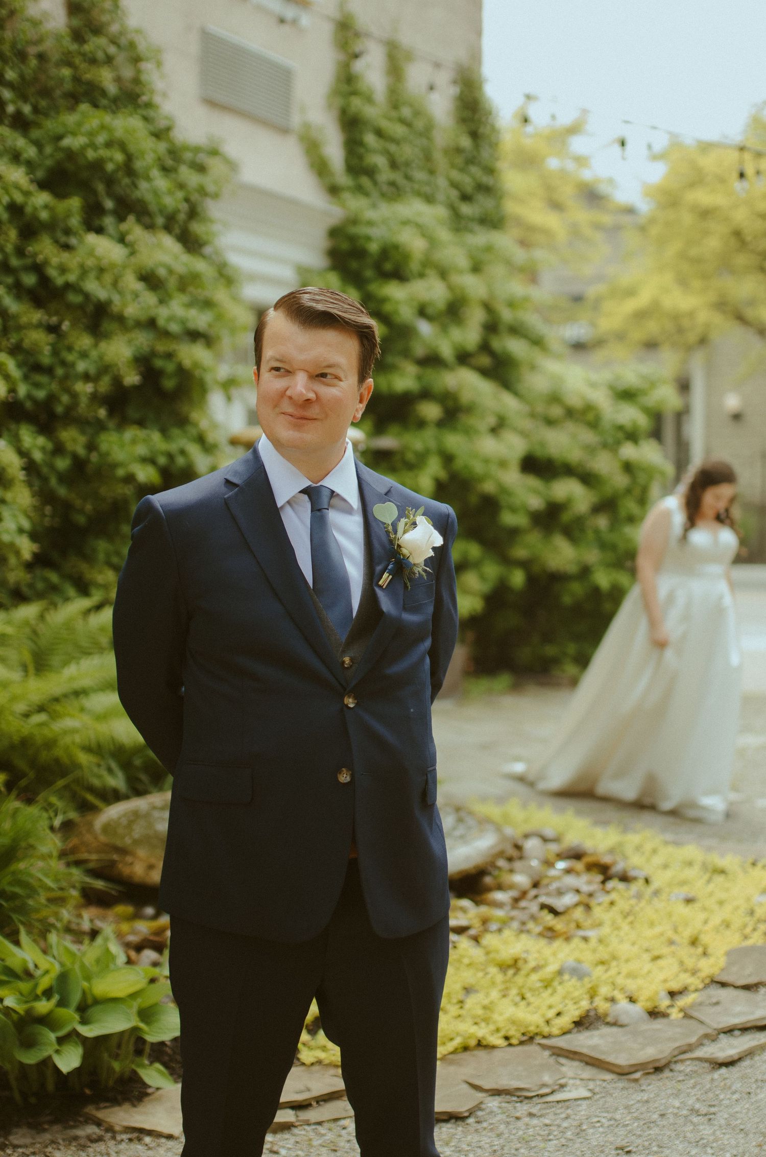 Groom in navy suit poses outdoors near lush greenery.
