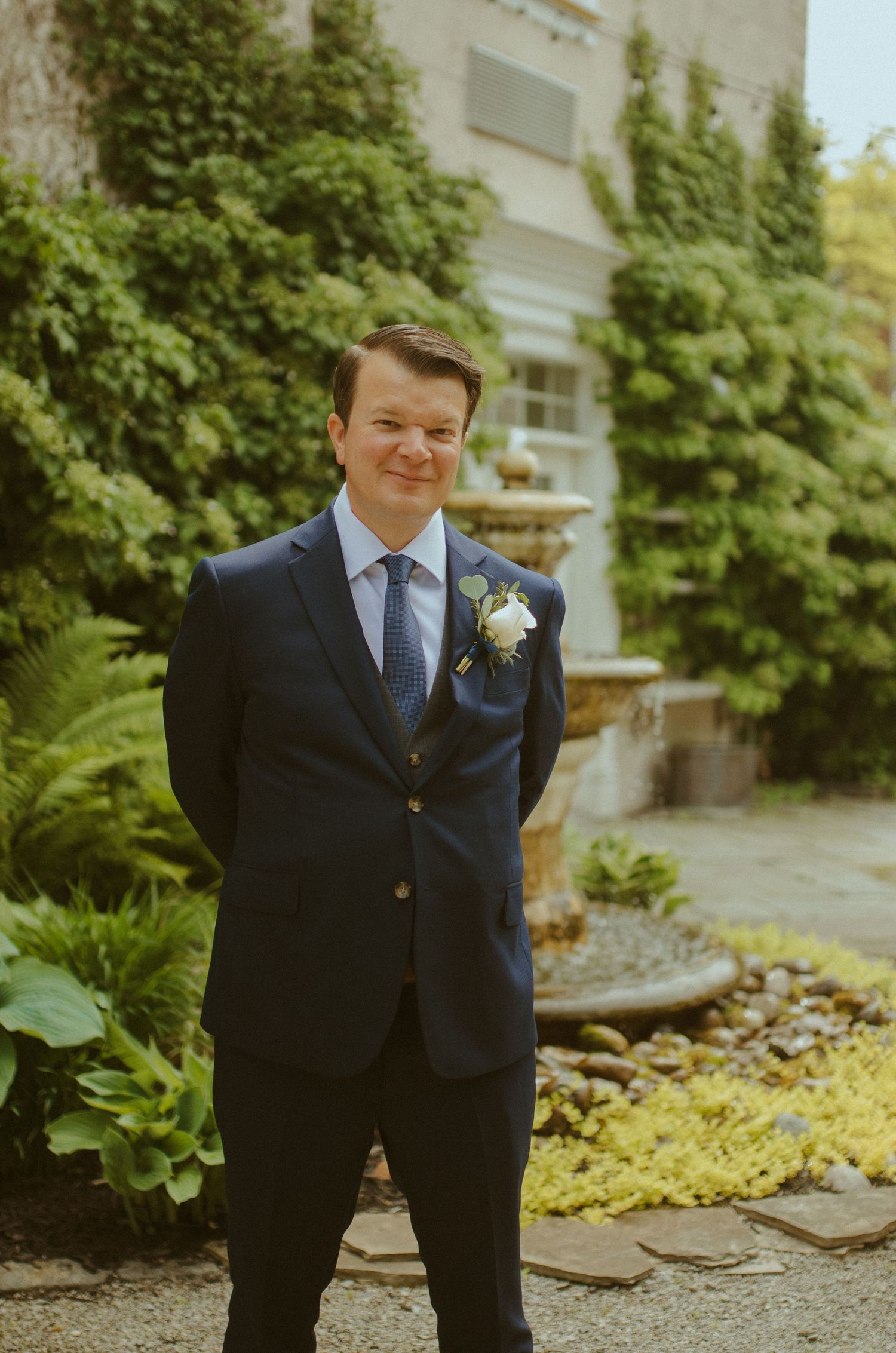 A groom in a navy suit poses in a garden setting with greenery in the background.