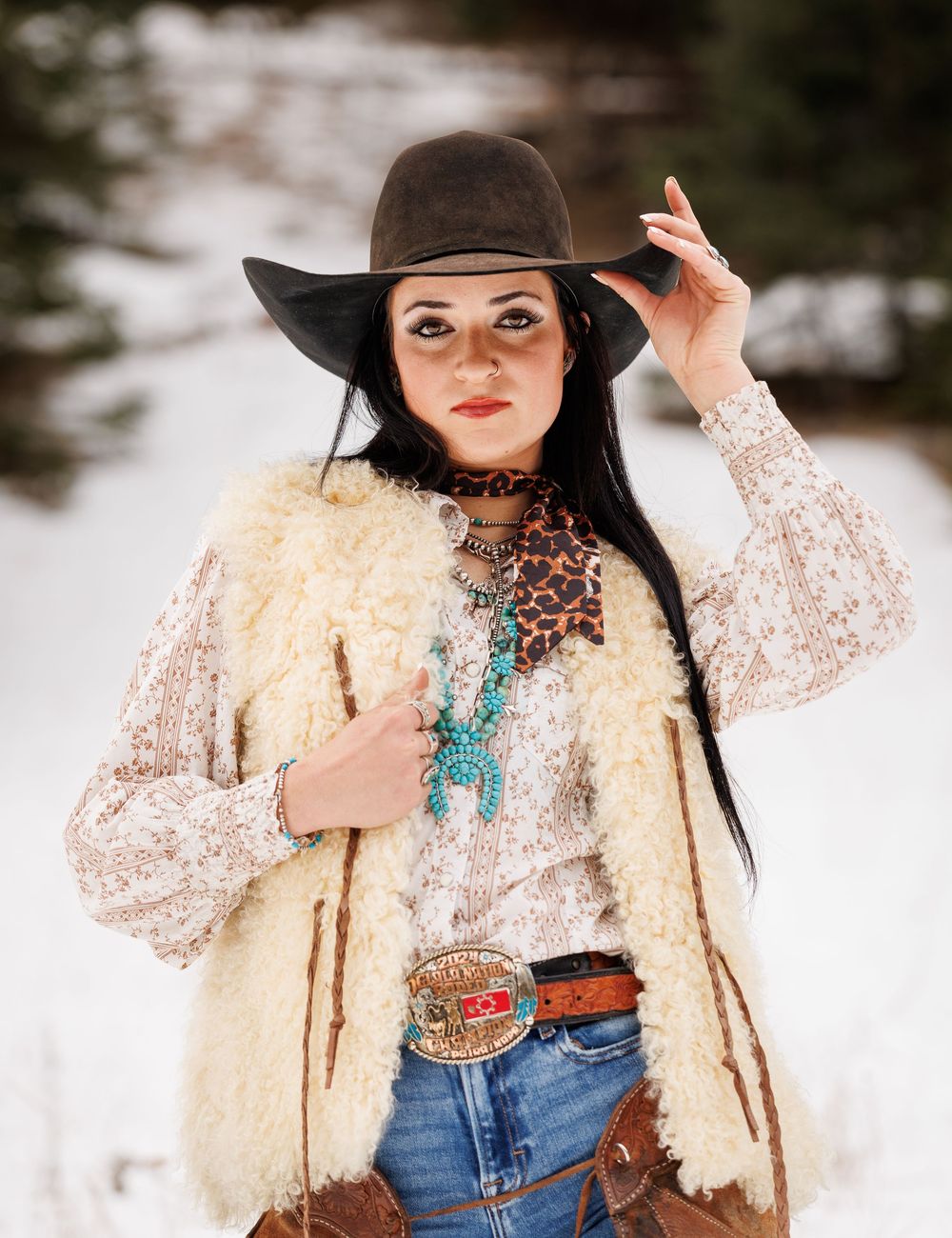 Western-inspired winter fashion with cream fur vest, lace top, turquoise jewelry, brown hat and denim jeans in snowy setting.