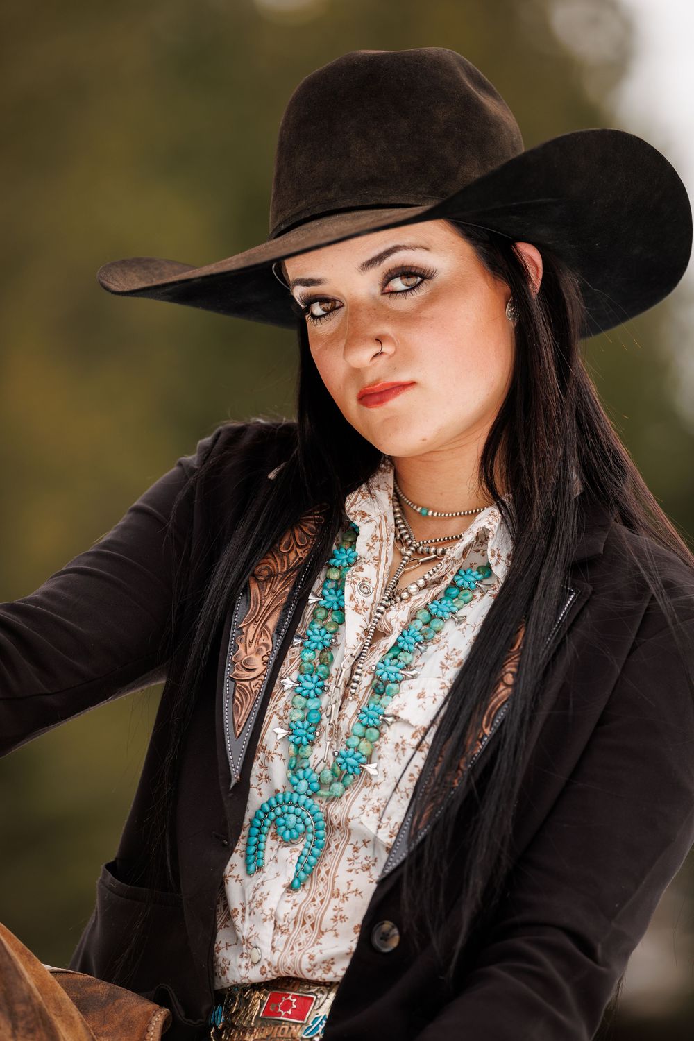 Western style portrait showing turquoise jewelry, black hat, and black jacket against a blurred outdoor background.