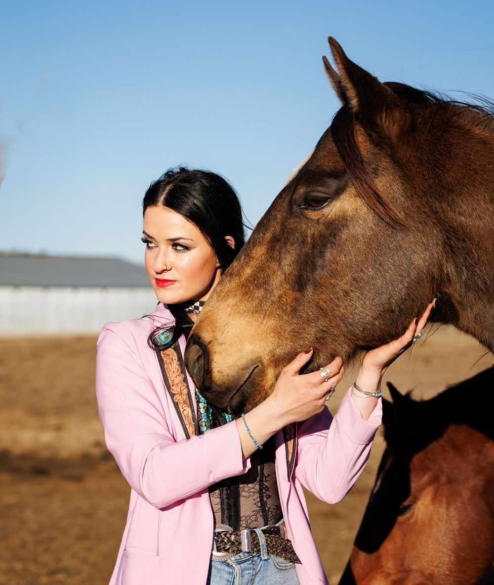 A person in a pink jacket shares a tender moment with a brown horse in an outdoor field setting.