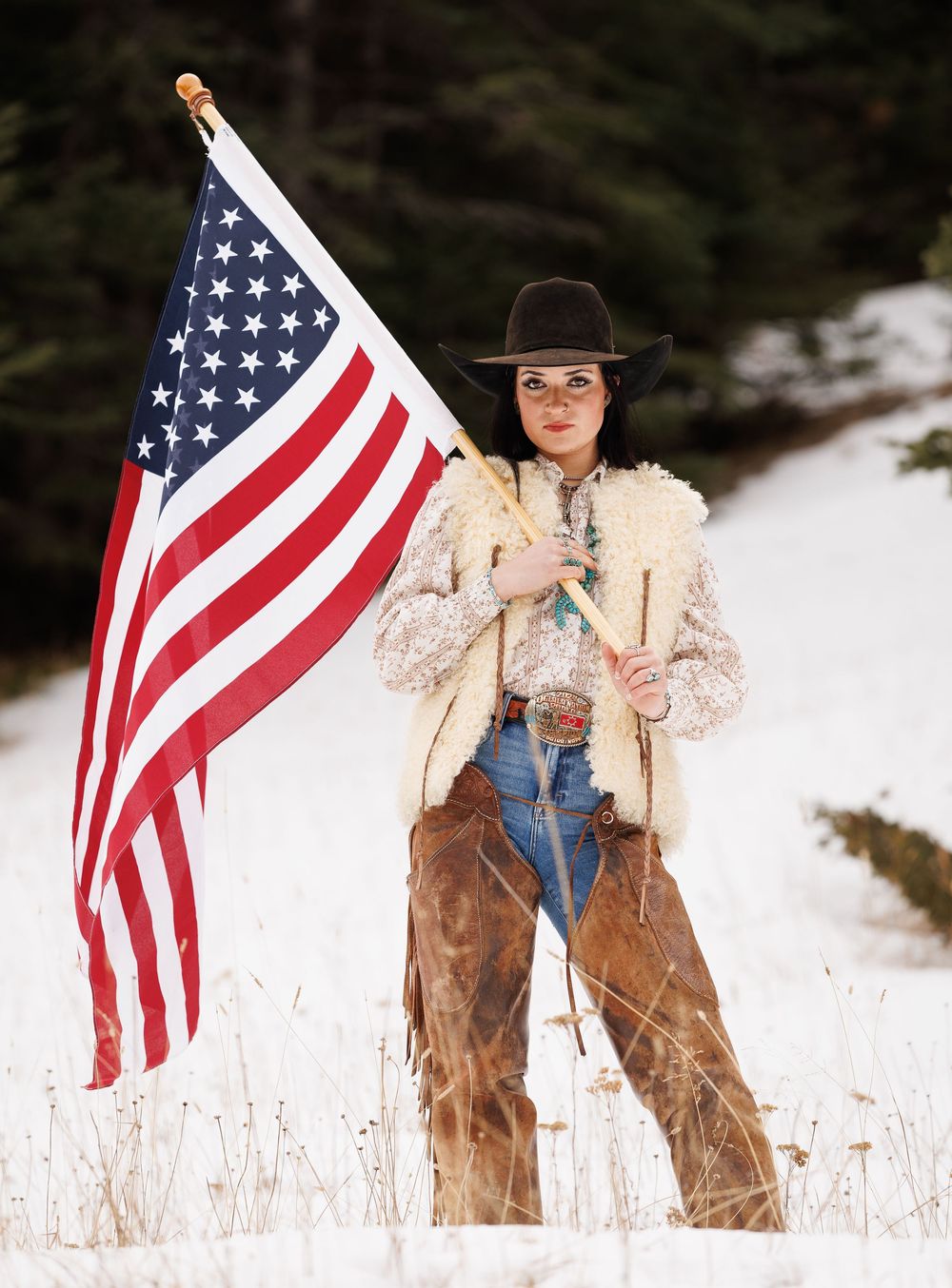 Person in western attire holding American flag in snowy field with rustic winter backdrop.
