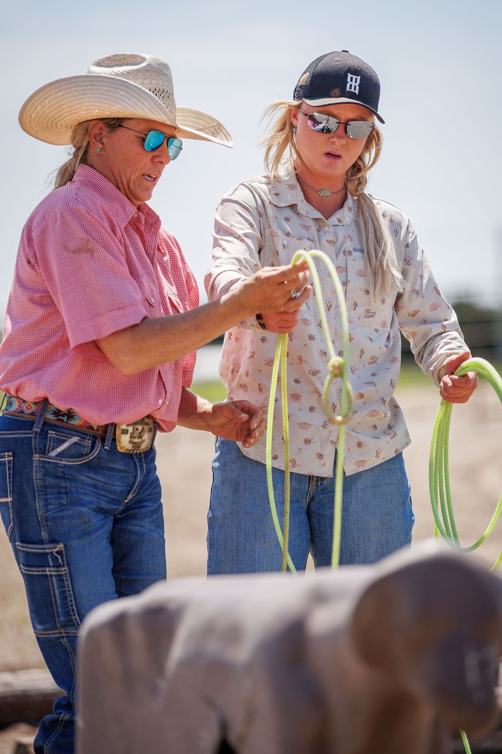 Two people in straw hats and pink shirts examine green onion stalks in a sunny agricultural field.