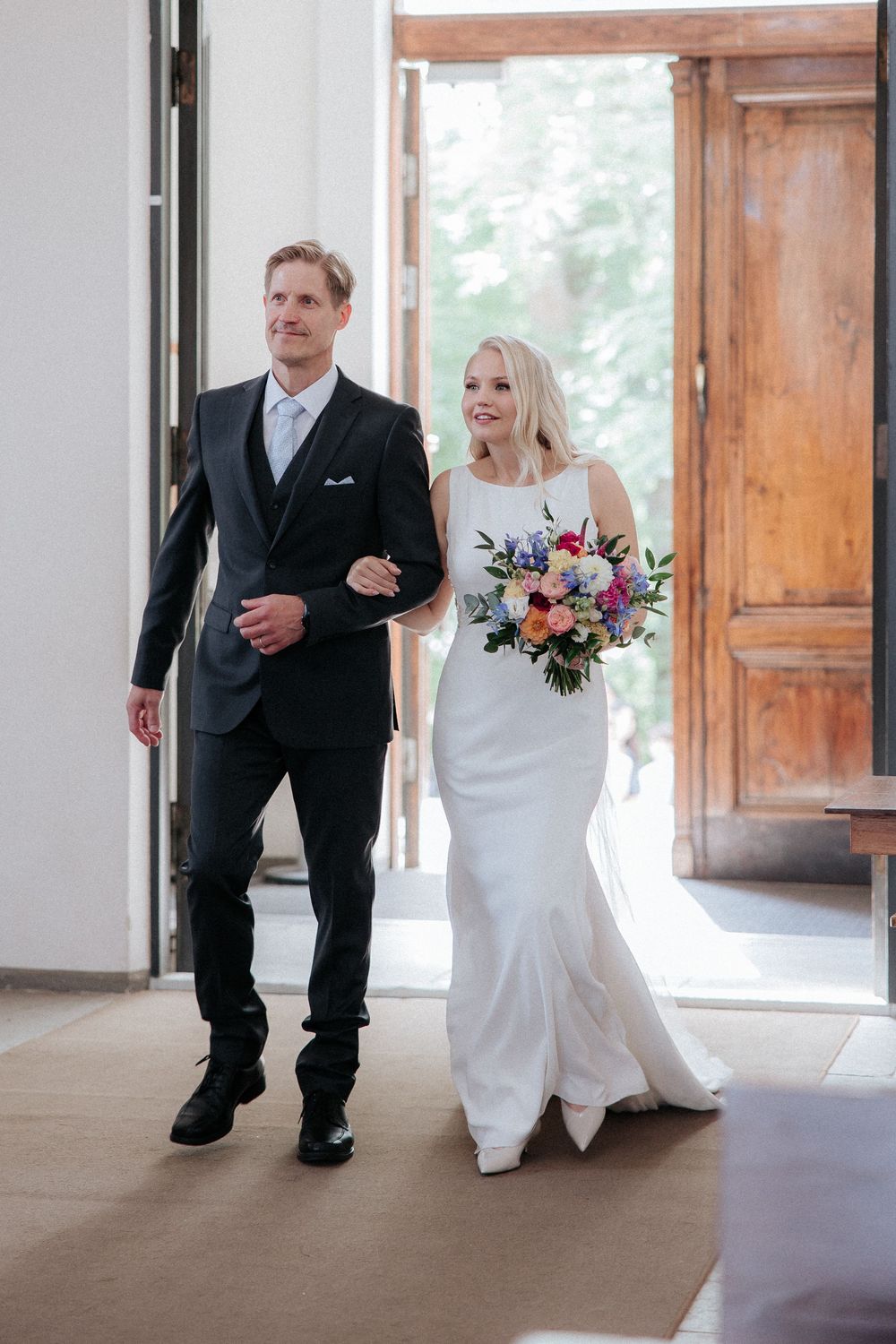 Wedding couple walks through doorway with bride holding colorful bouquet while wearing elegant white gown.