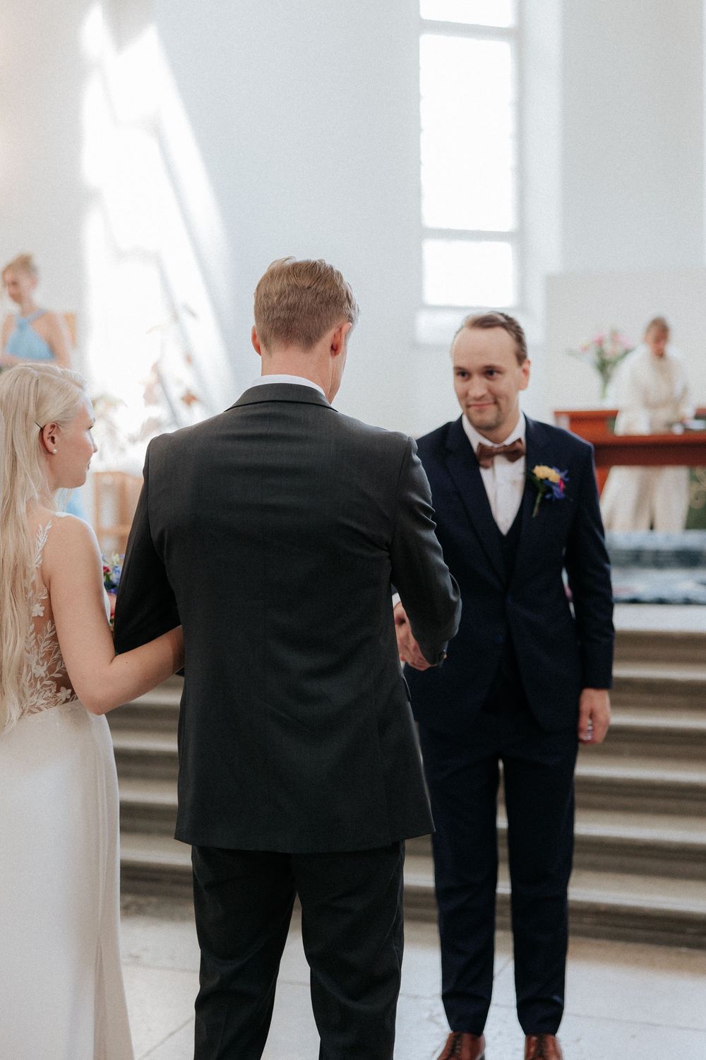 A wedding ceremony at a church with a couple and officiant standing at the altar steps exchanging meaningful gestures.