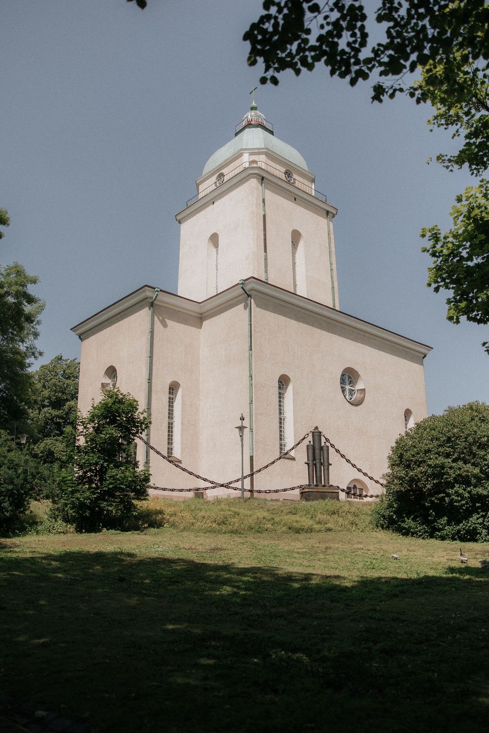 A white church with a domed tower sits atop a grassy hill against a blue sky with trees in the foreground.