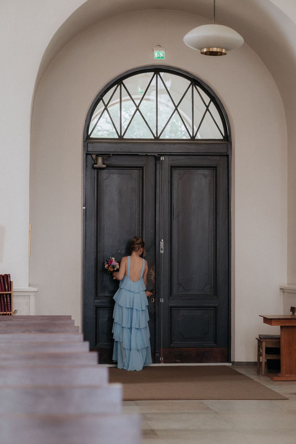 Person in light blue tiered dress stands before tall dark doors with arched geometric window in elegant hallway.