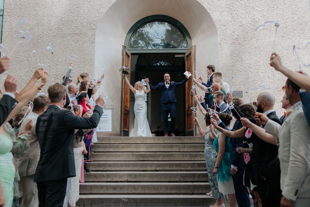A group of wedding guests stand on stone steps outside a church entrance throwing petals at the departing couple.
