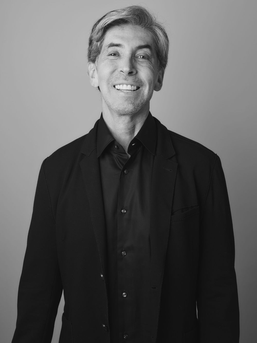 Professional black and white portrait of a smiling person in dark business attire against a light background.