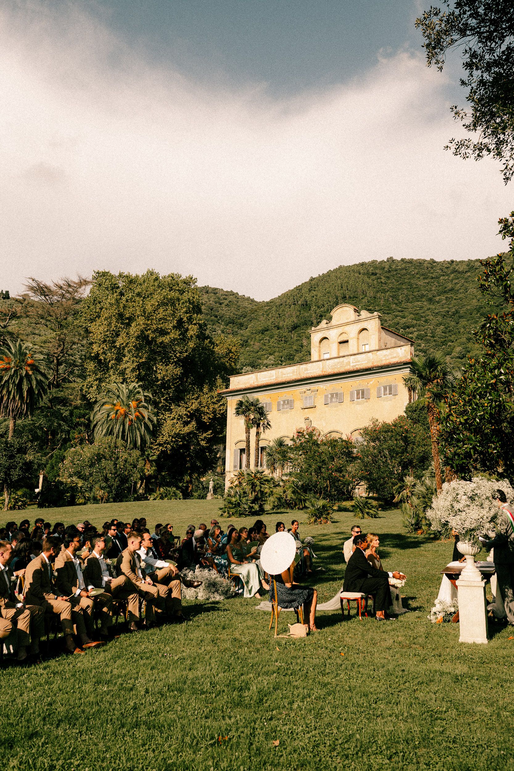 People gather on a grassy hillside for an outdoor event with a historic mansion and mountains in the background.