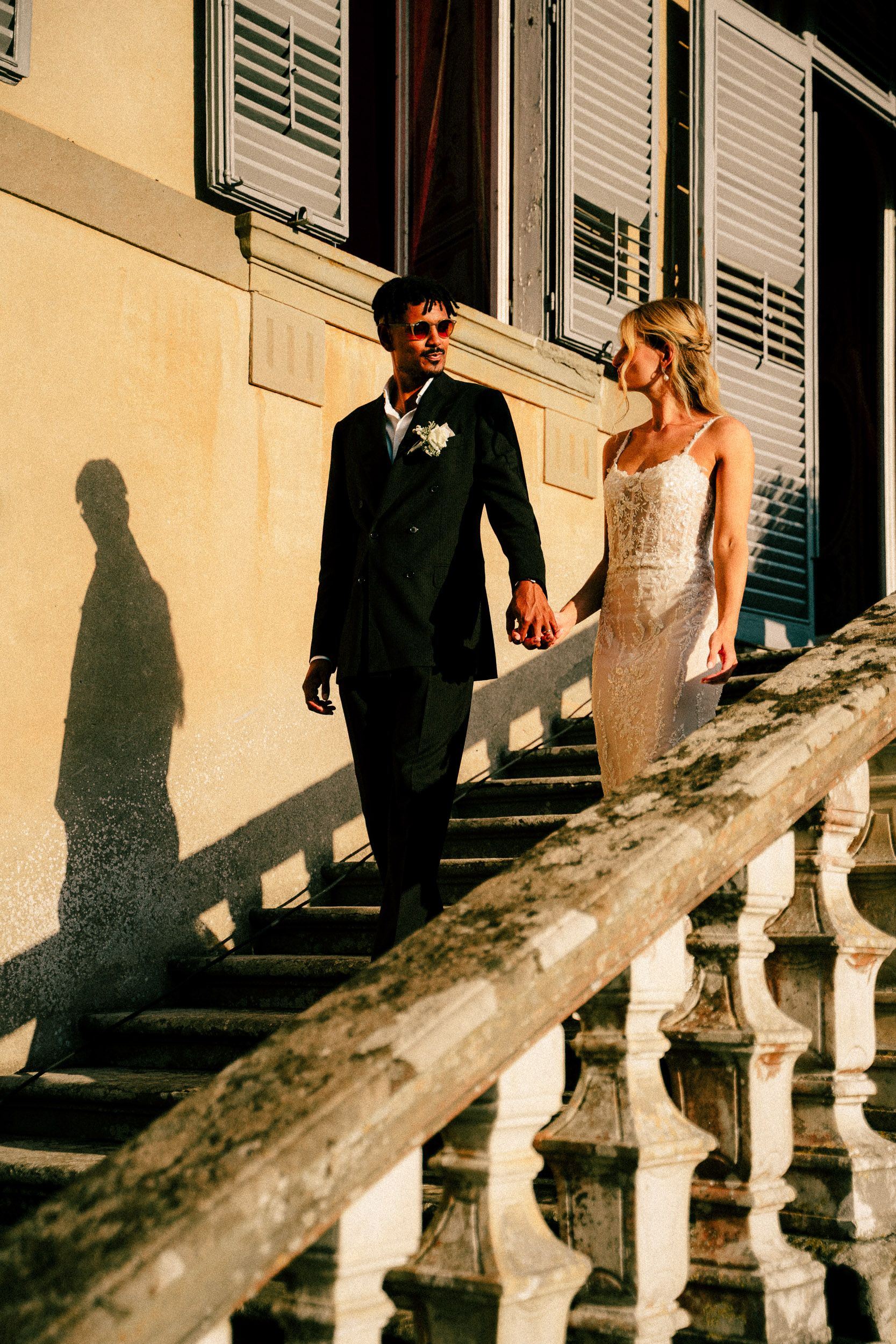Elegant couple in wedding attire walking together on a sunlit stairway with dramatic shadows cast on the wall.