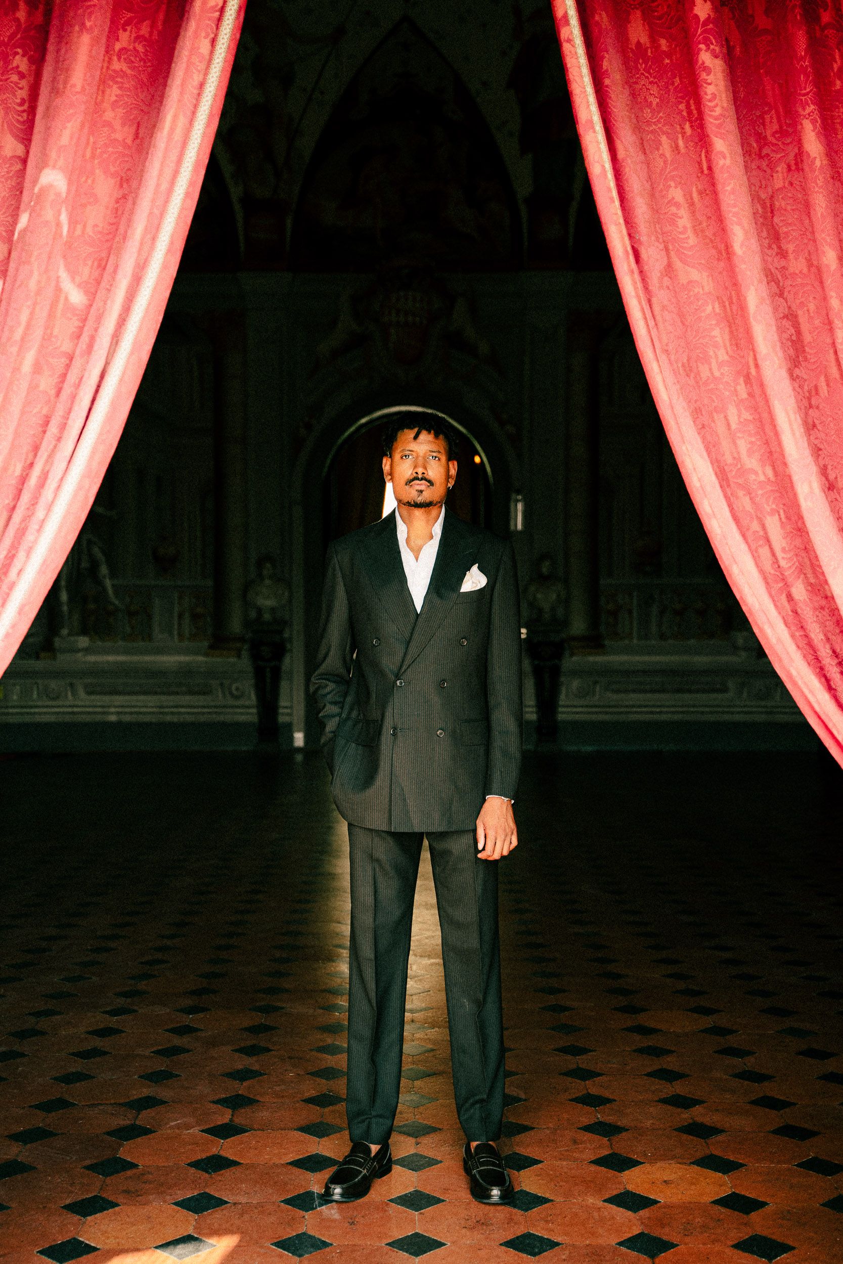 A person in a suit stands in front of red theater curtains on a black and white checkered floor.
