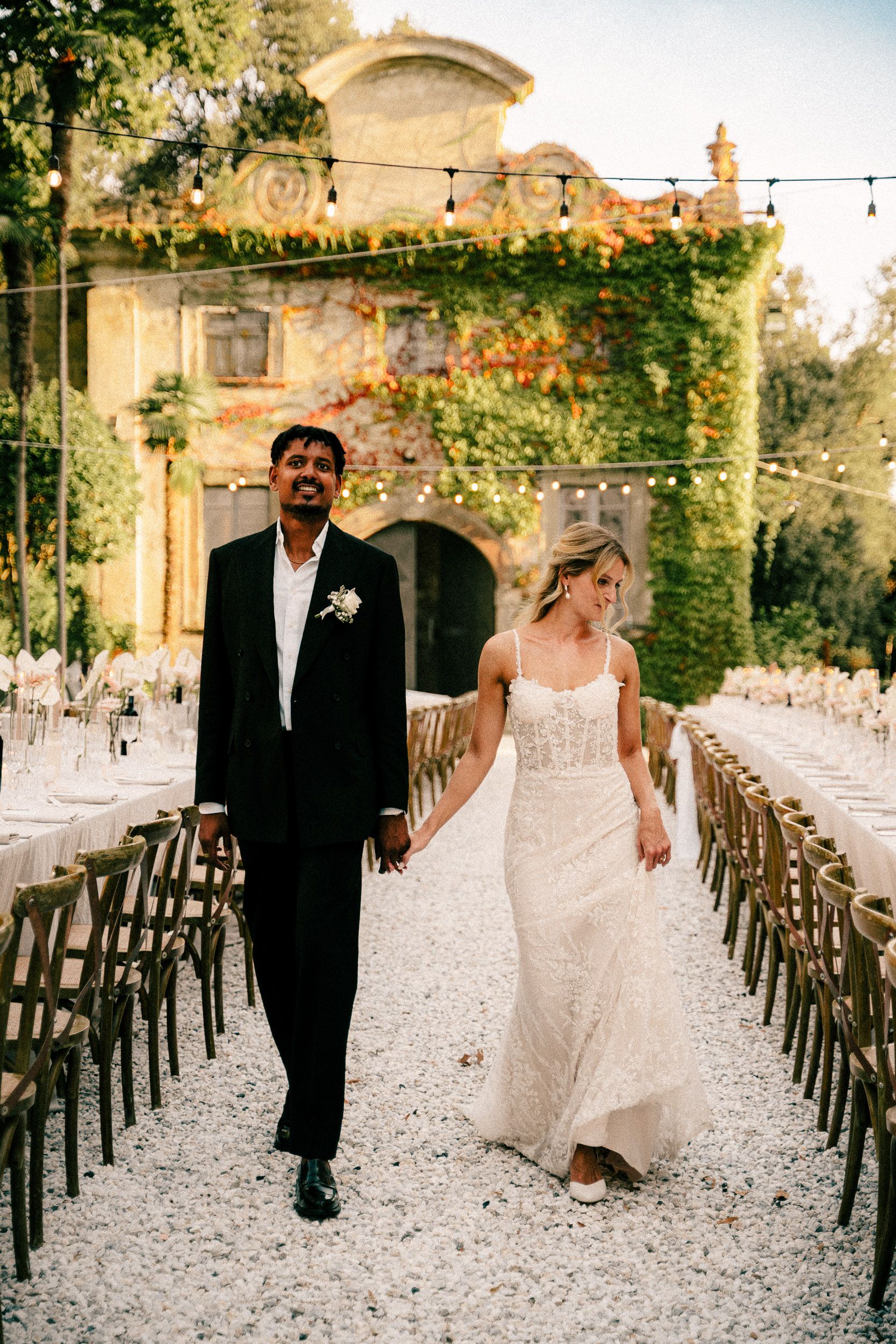 A couple walks through an elegant courtyard ceremony setup with rustic wooden chairs and string lights at sunset.