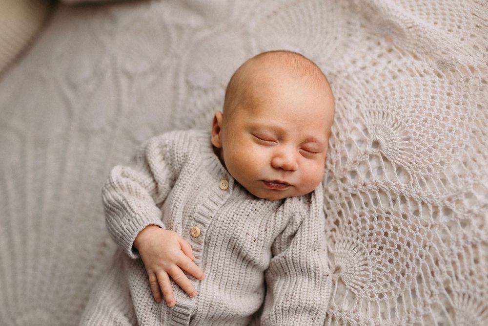 A sleeping baby dressed in a knitted gray sweater lies peacefully on a soft textured blanket.