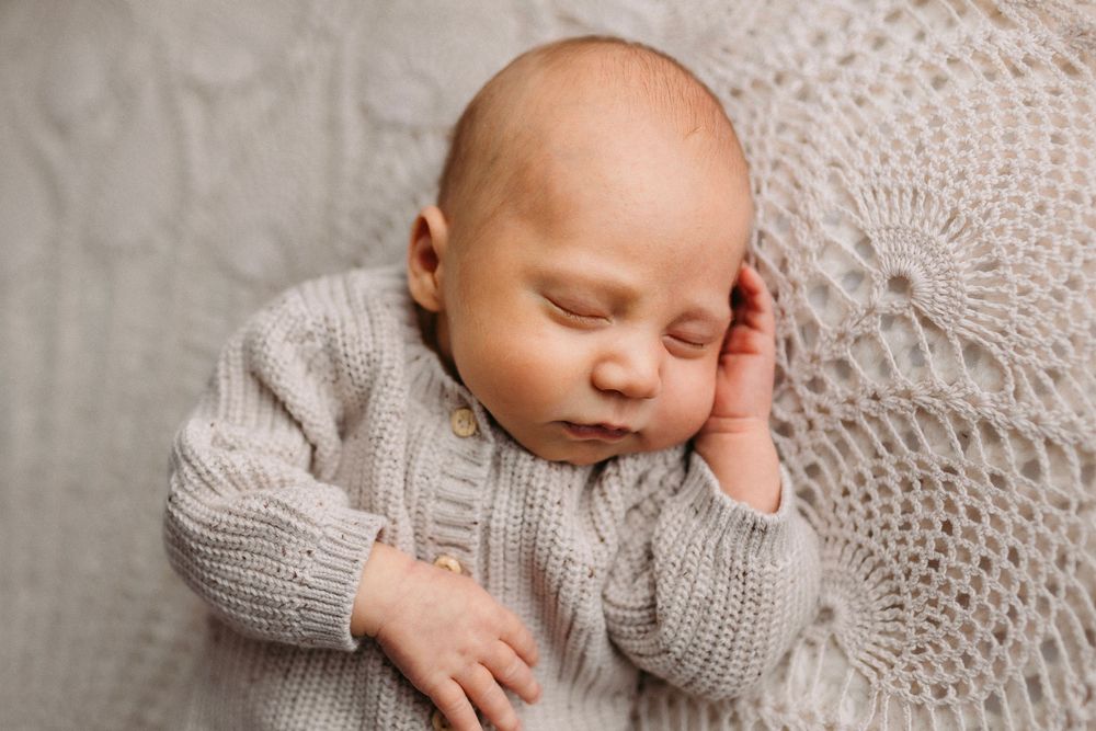 Newborn baby wearing gray knit sweater sleeps peacefully on soft textured blanket.