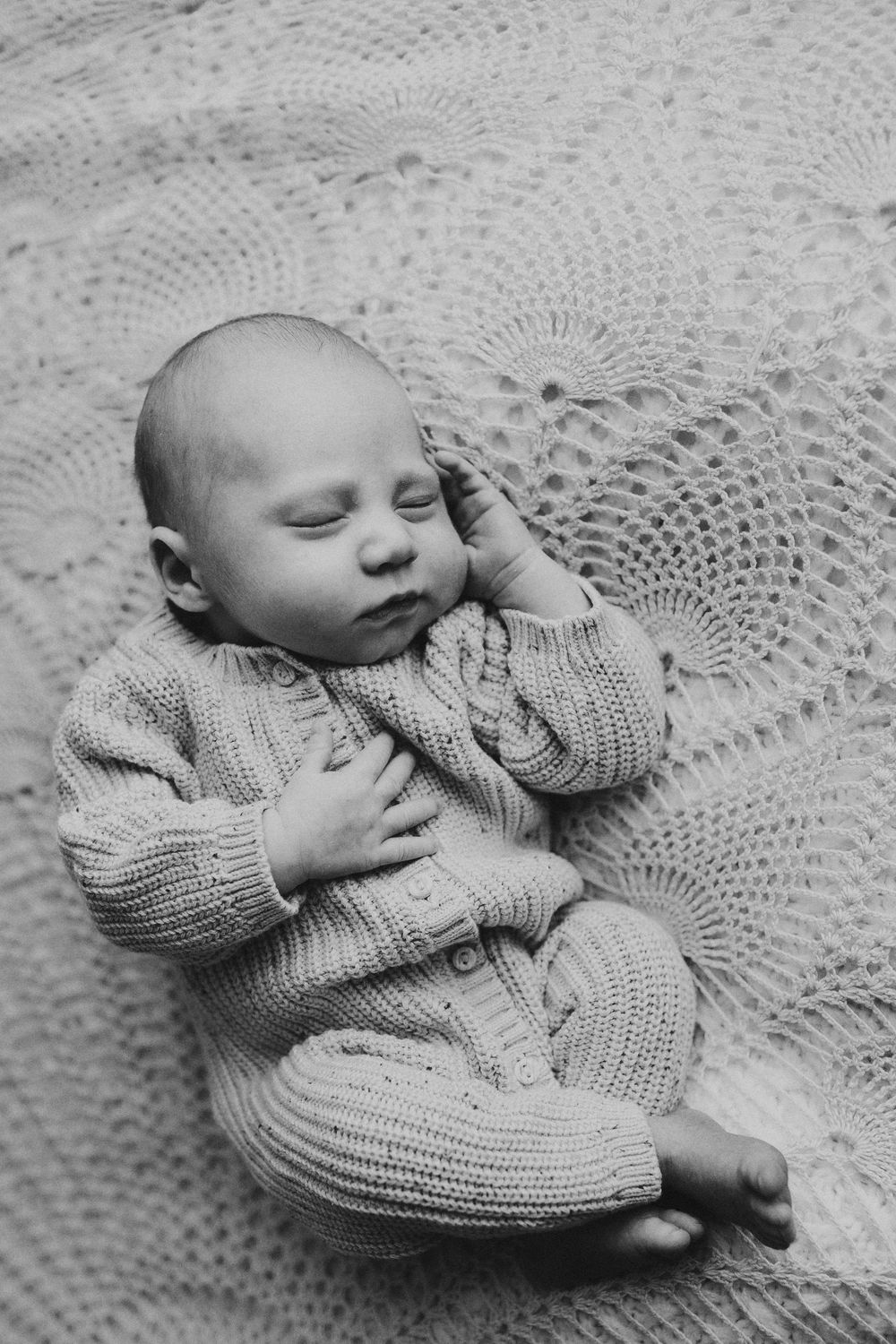 Sleeping newborn wearing gray knit sweater lying on lace blanket in peaceful pose.
