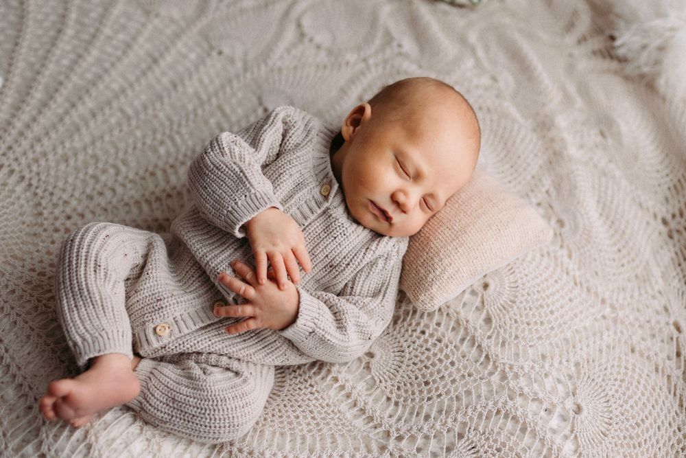 Newborn in cozy knit sweater resting peacefully on textured white blanket during photo session.