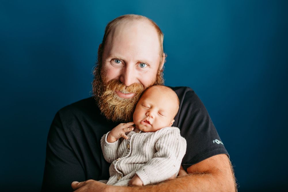A person with a ginger beard sits in front of a dark blue background wearing a black shirt and looking thoughtful.