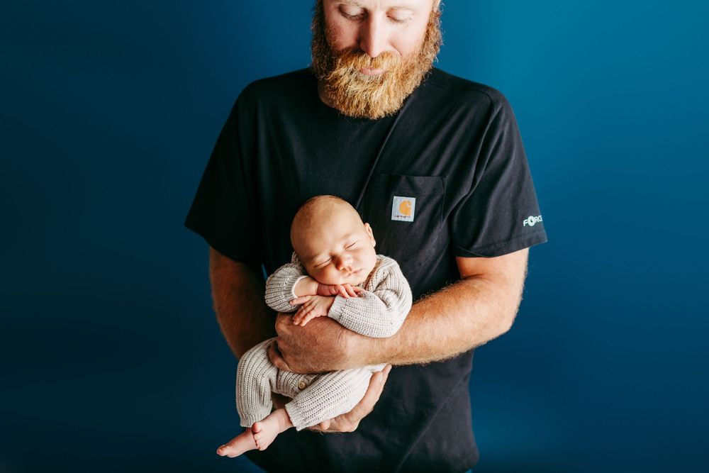 A parent wearing a black t-shirt cradles and soothes a sleeping newborn baby against a blue background.