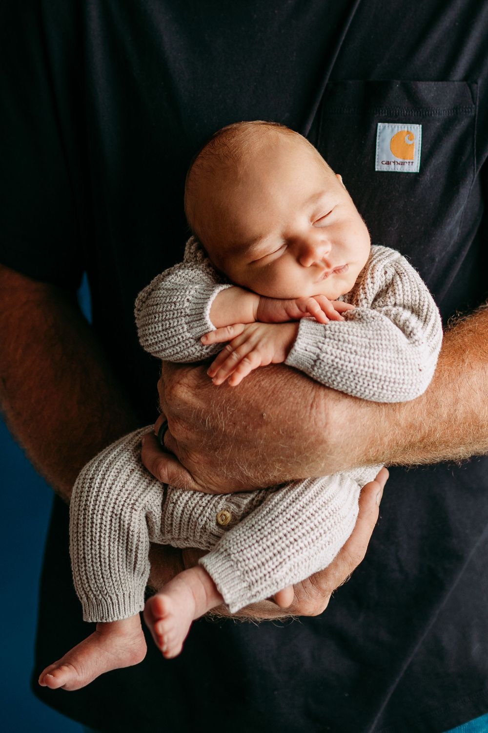 A sleeping newborn baby wrapped in a cozy gray knit blanket being cradled in strong arms against a dark background.