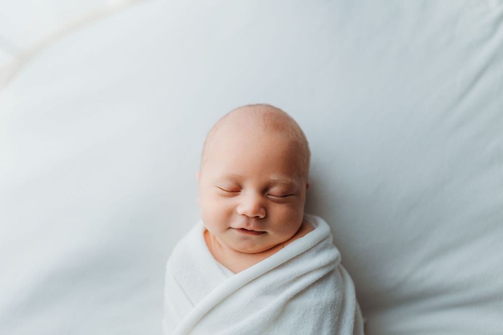 Sleeping newborn baby in white wrap positioned on soft light bedding during professional photography session.