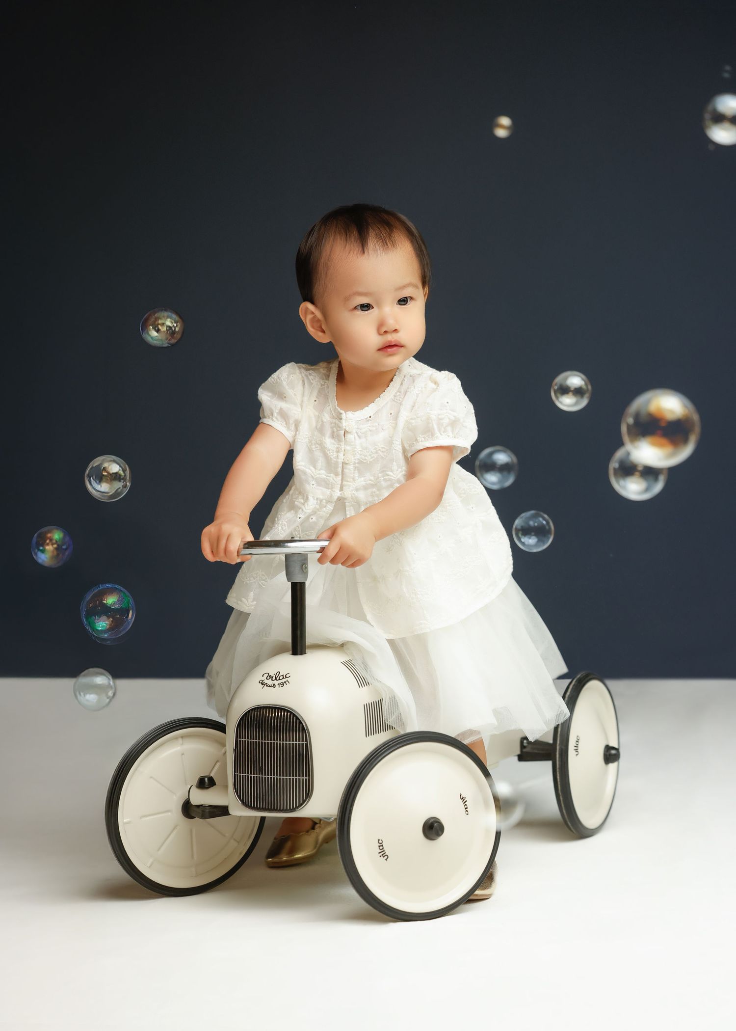 Child in white dress rides vintage-style white pedal car while soap bubbles float through dark studio setting.