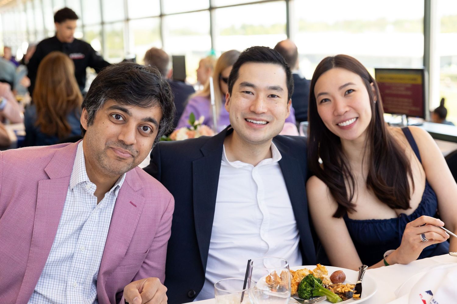 Three smiling people, two men and a woman, posing together at a dining event with food on the table.