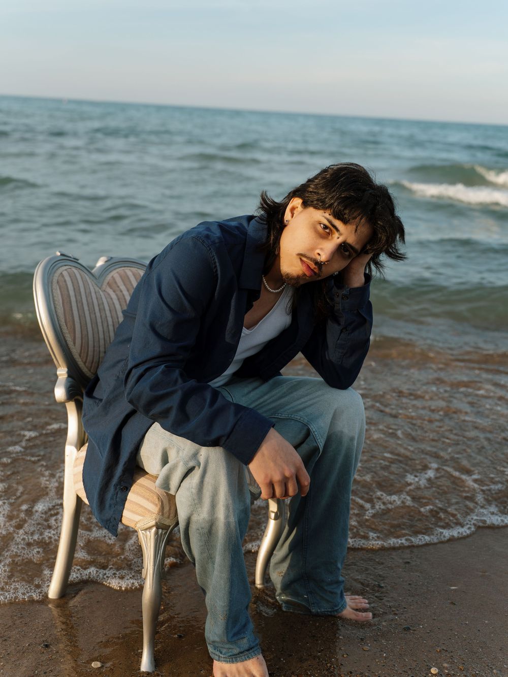 Man in dark blue button up, white t-shirt, and blue jeans poses on a tan, heart shaped chair on the beach.