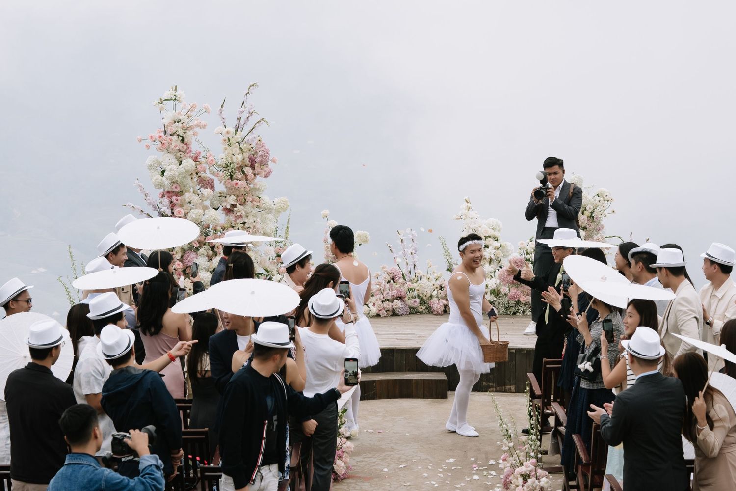 Outdoor wedding ceremony on a misty cliffside with white floral arch and guests holding parasols.