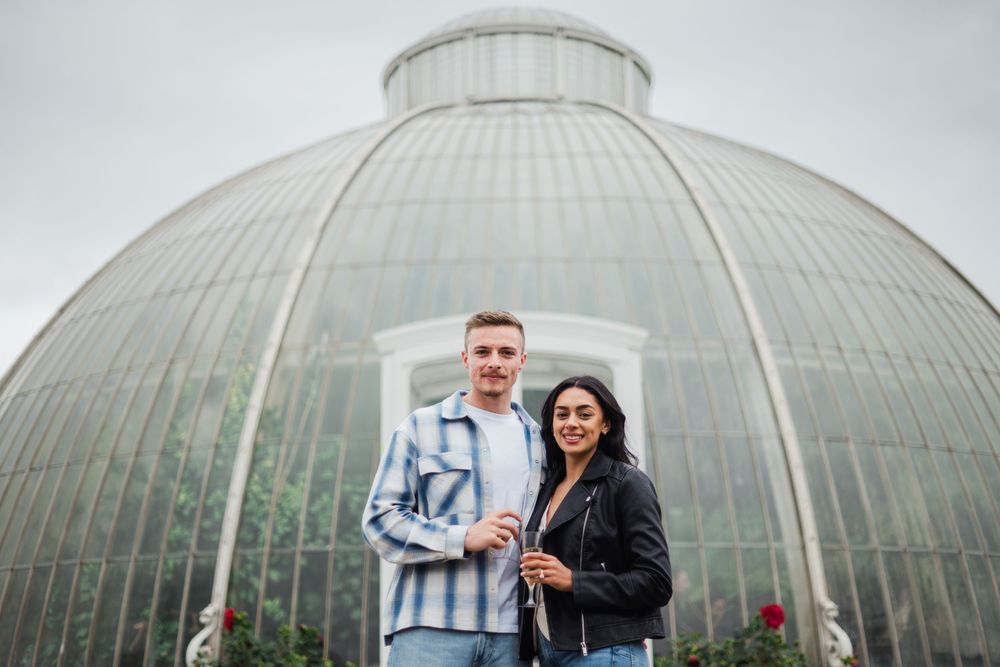 A couple poses for an engagement photo shoot in front of a large glass dome greenhouse structure.