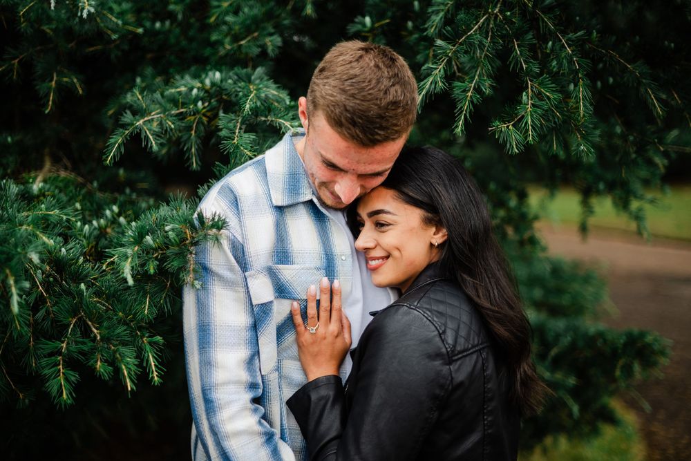 A couple shares a tender moment while embracing near evergreen branches in an outdoor setting.