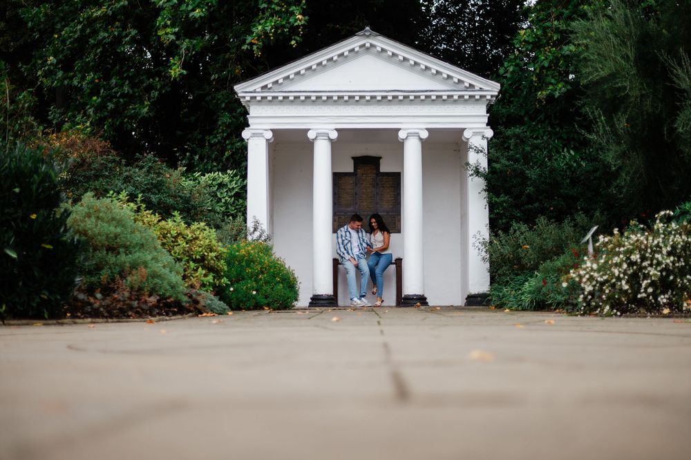 A couple stands together in a classical white gazebo with columns surrounded by garden landscaping.