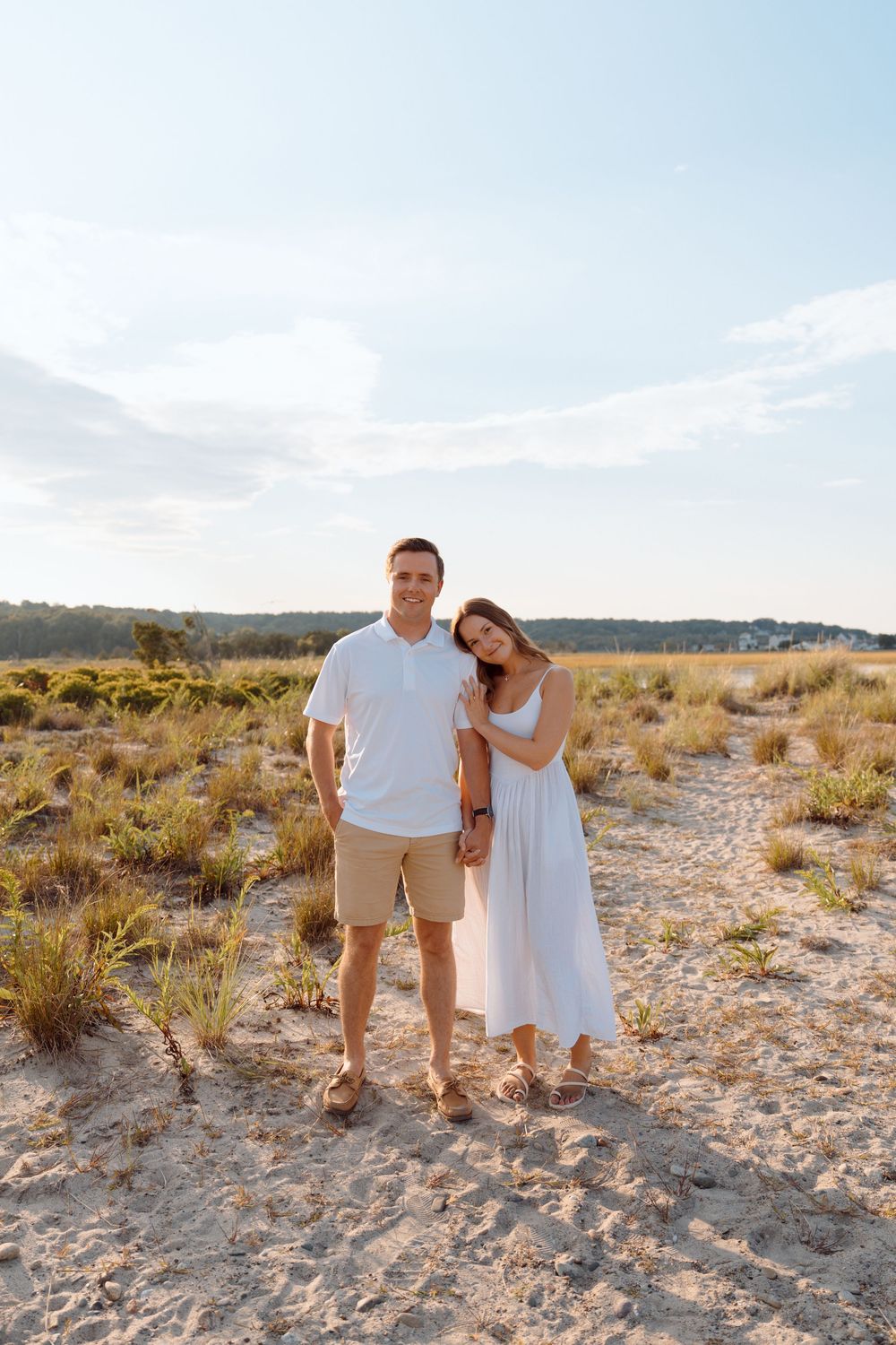 A couple walking together along a sandy beach path at sunset in casual white summer attire.