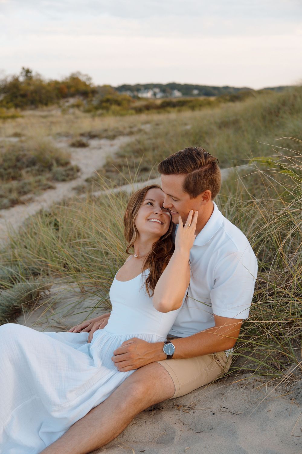 A romantic couple embraces on sandy dunes at sunset wearing white and beige casual attire.