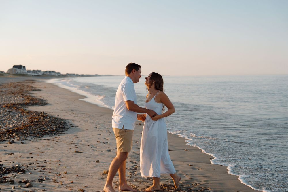 A romantic couple embraces on a sandy beach at sunset with ocean waves gently rolling in the background.