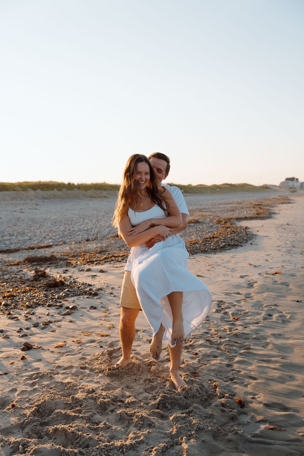Playful beach photoshoot with couple in white dress and khaki shorts on sandy shoreline.