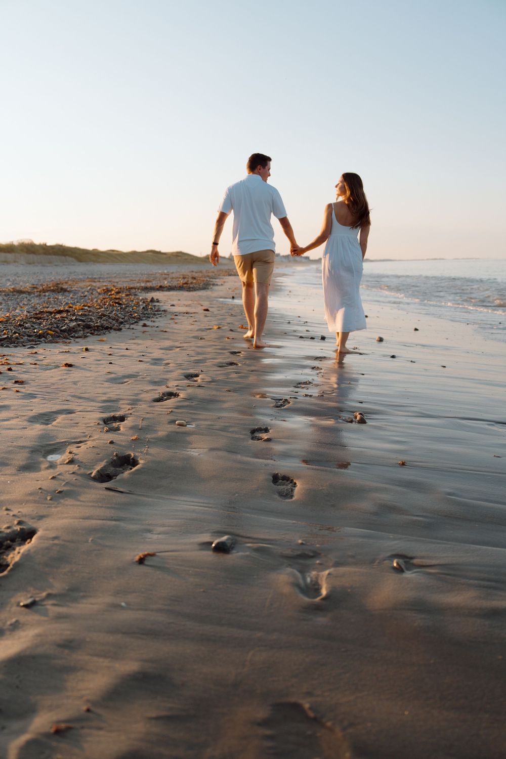 A couple walks along a sandy beach at sunset leaving footprints in the wet sand as waves gently lap the shore.