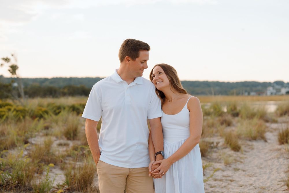 A couple shares a romantic moment while walking along a sandy beach path at sunset wearing white and khaki clothing.