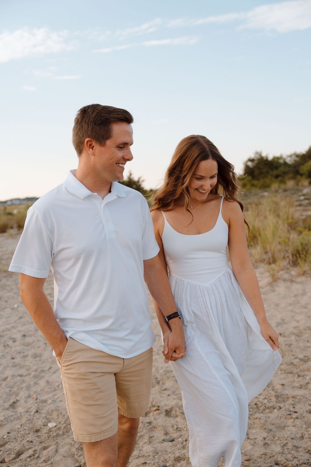 Couple walking hand in hand on sandy beach path at sunset wearing white summer outfits.