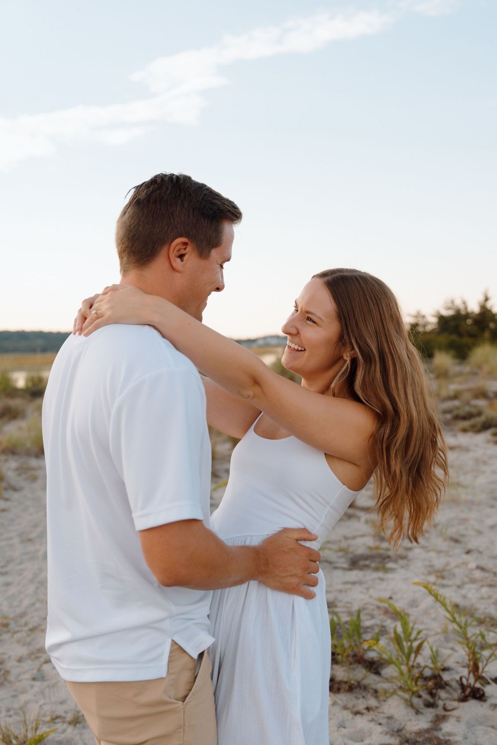 A couple in white outfits embracing and smiling at each other on a beach during sunset.