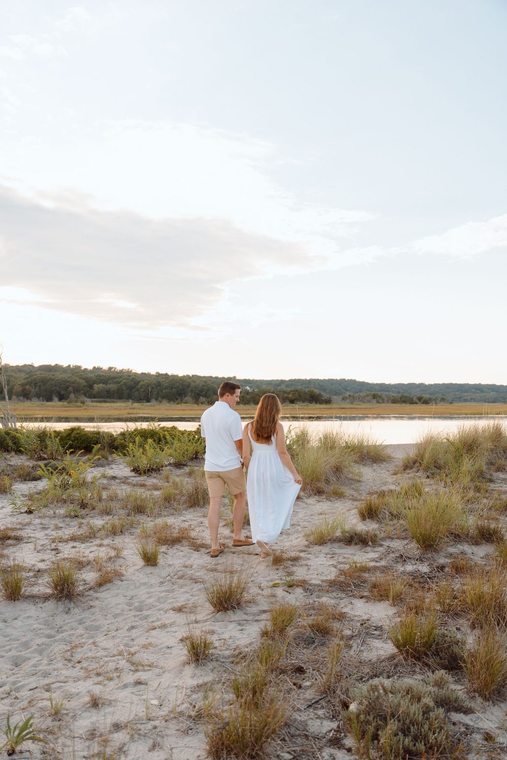 Couple walking together at sunset on a sandy beach with coastal grasses and dunes in the background.