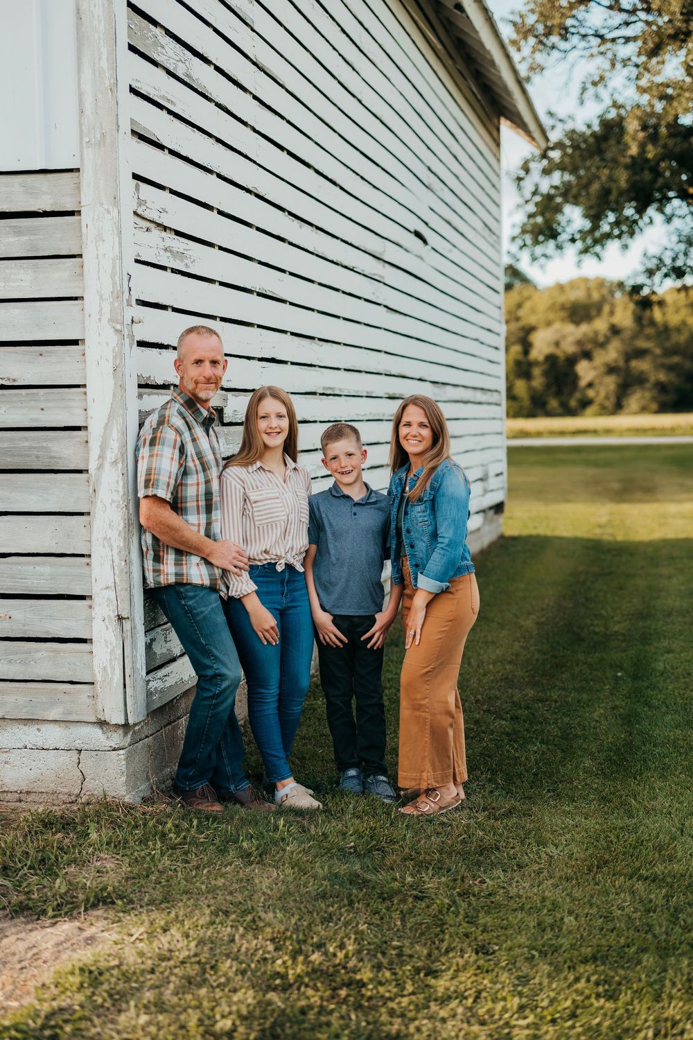 Family poses casually against white farmhouse wall in casual attire during golden hour outdoor photoshoot.