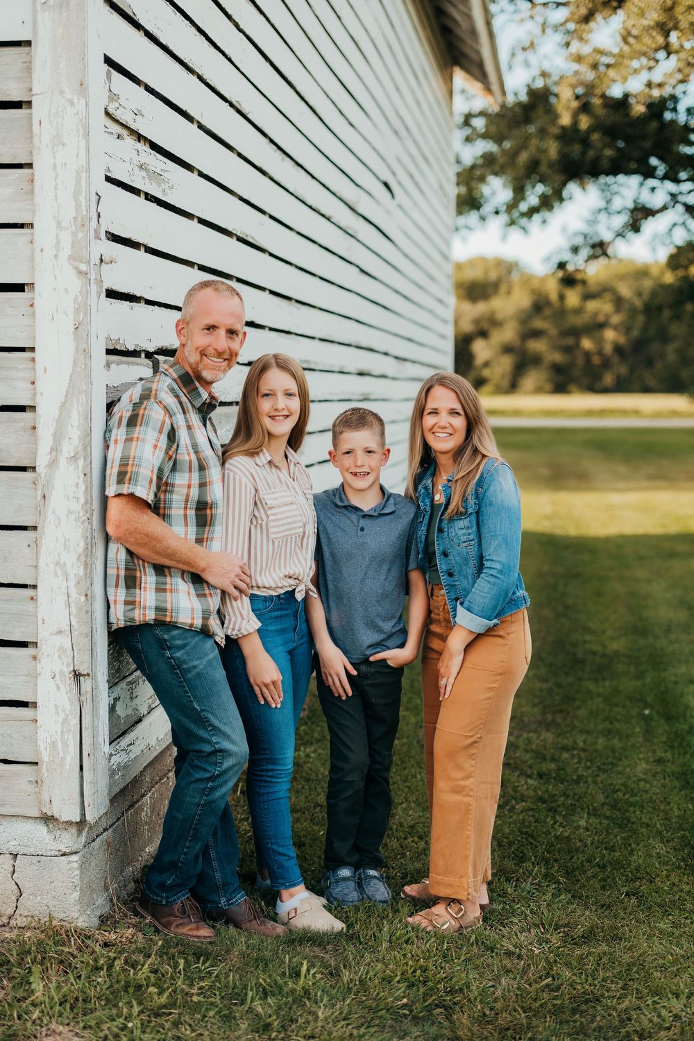 A group poses casually against a rustic white barn wall in coordinated casual outfits during golden hour.