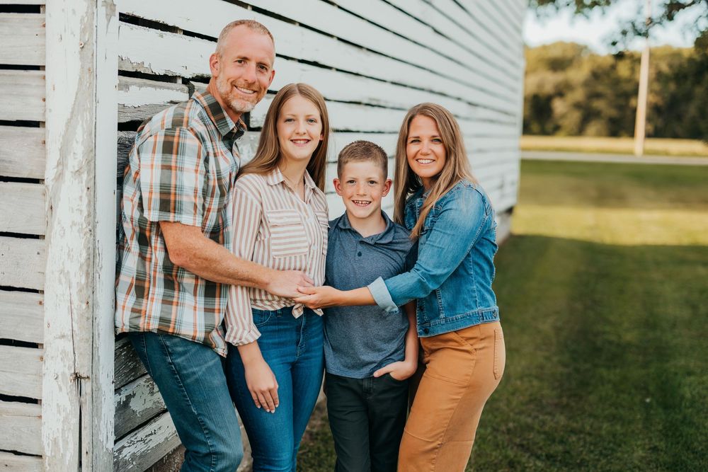A loving family poses together in coordinating outfits against a rustic white wood barn paneled backdrop outdoors.