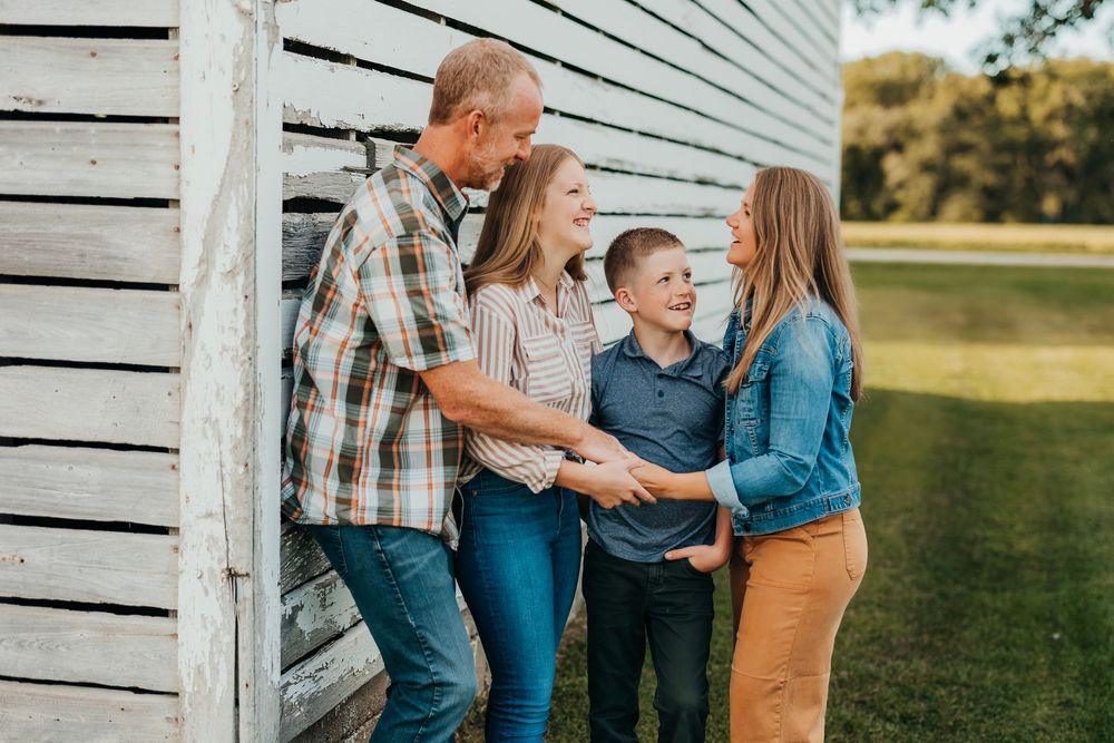 A happy family of four shares a candid moment together while leaning against a rustic white barn wall.