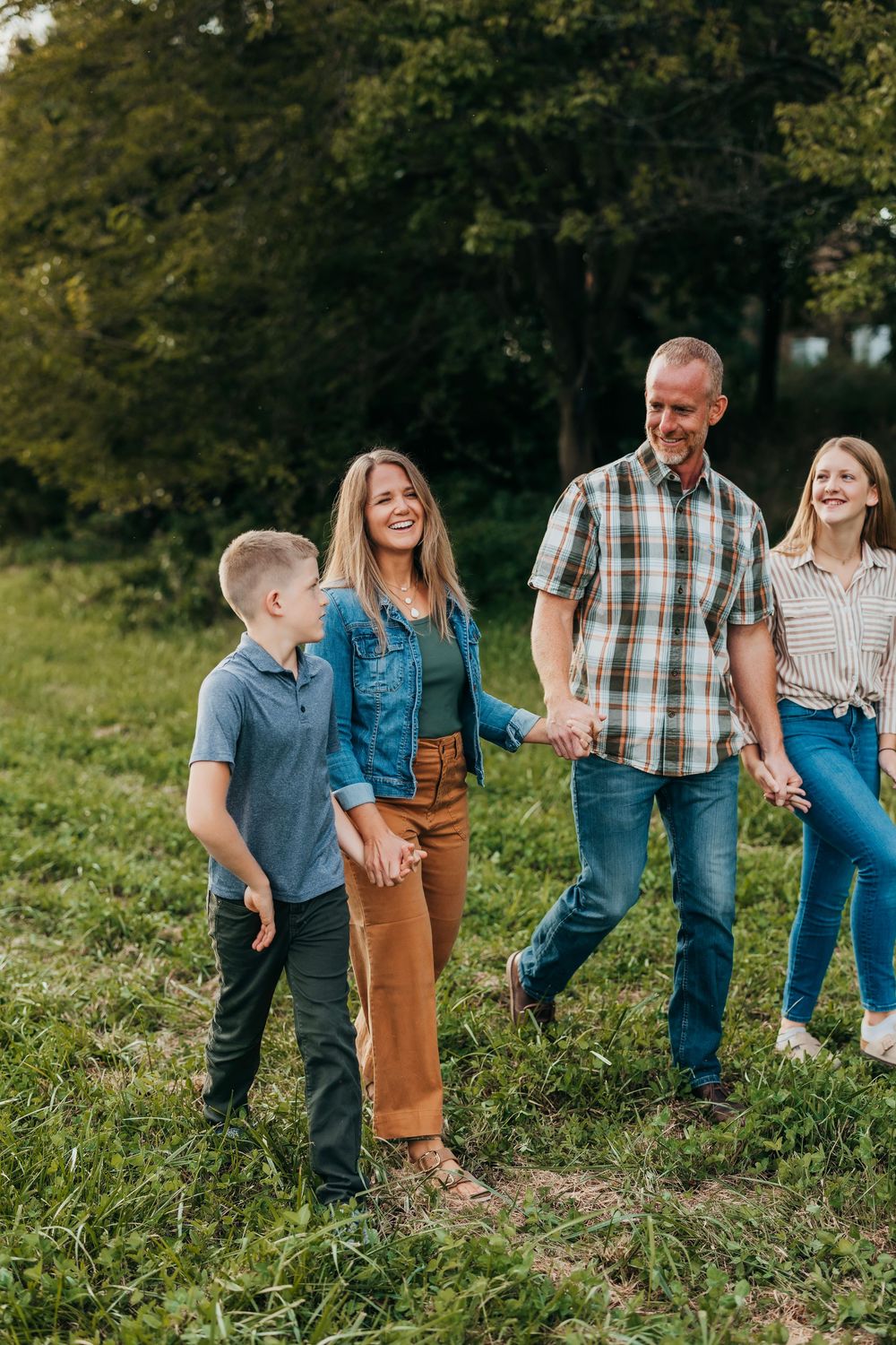 Family in casual denim and plaid attire poses together on grassy hillside during outdoor photo session.