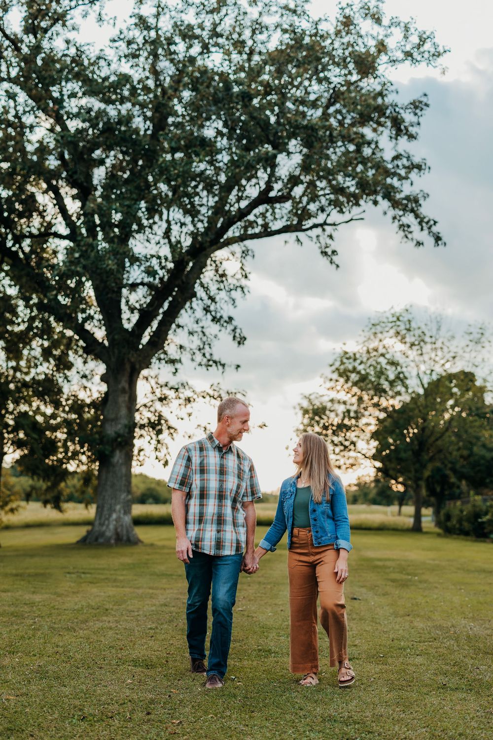 Family walks together across grassy field with large tree and sunset sky in background.