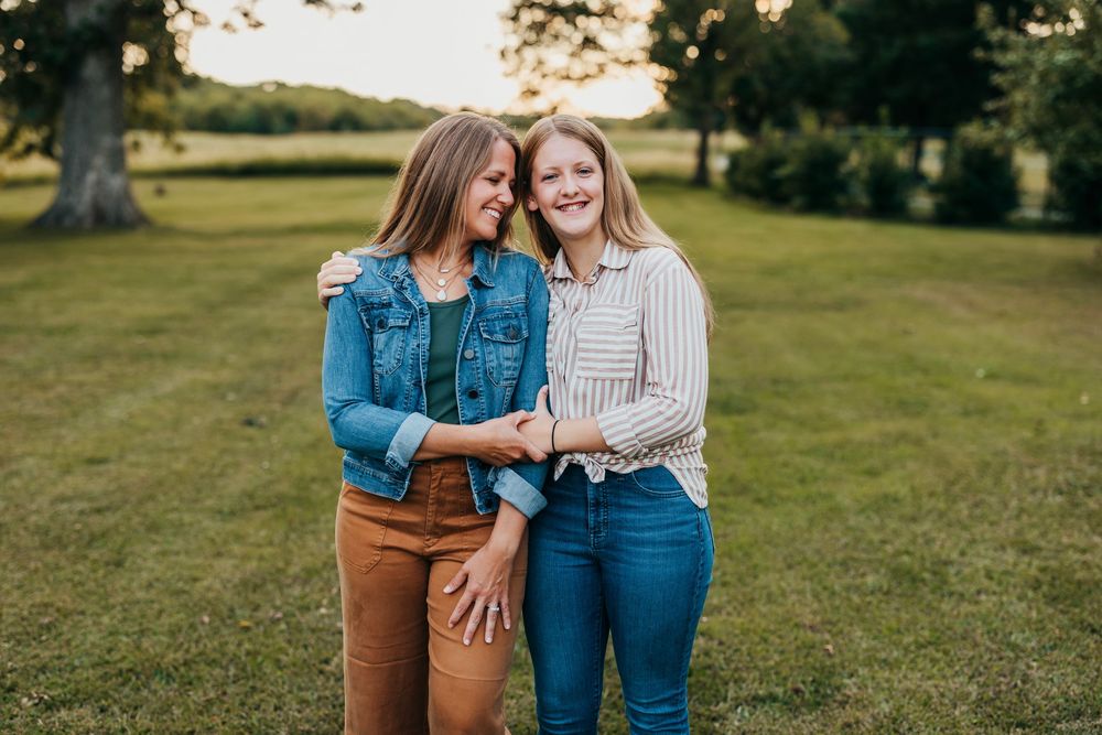 Two people in casual denim and knit outfits share a candid moment outdoors in a grassy field at sunset.