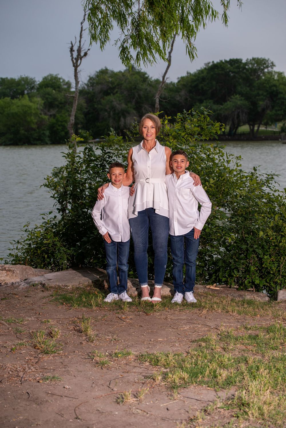Three people in matching white shirts and jeans pose together by a lake with trees and greenery in the background.
